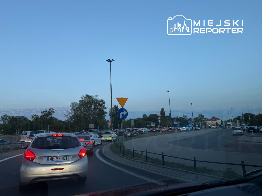Ruch samochodowy w korku na rondzie, z widocznymi pojazdami i sygnalizacją świetlną w tle.
