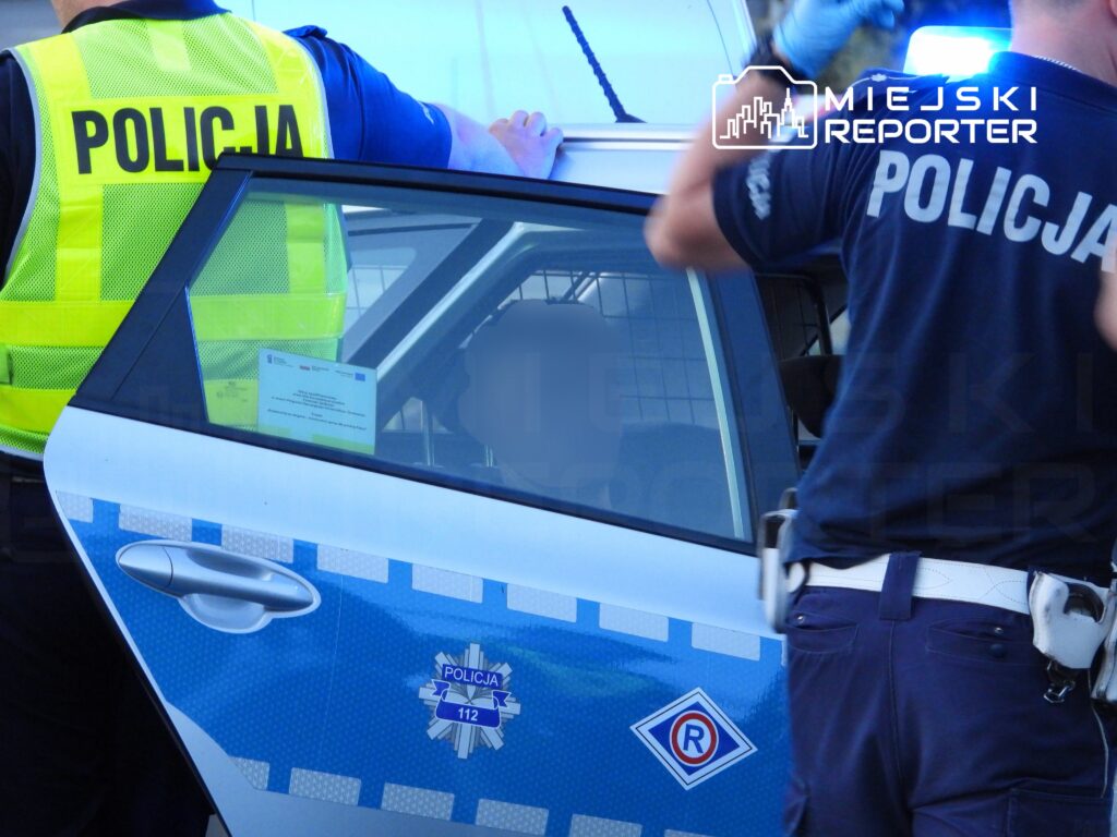 Dwaj policjanci w mundurach, jeden z nich w kamizelce odblaskowej, interweniują przy policyjnym samochodzie.