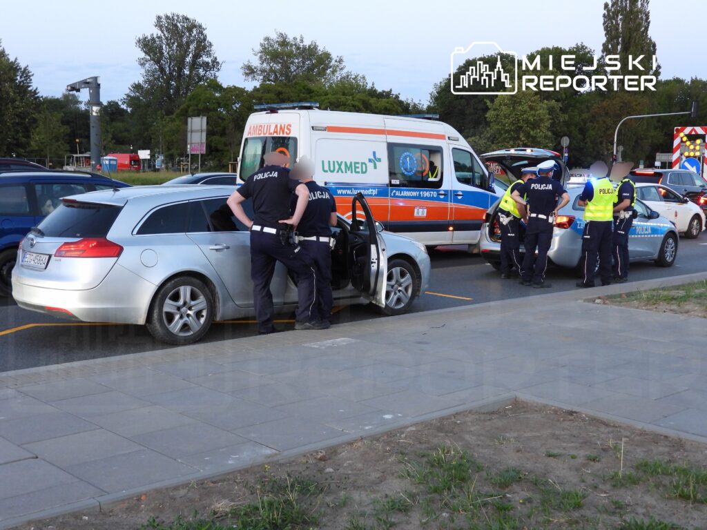 Grupa policjantów prowadzi czynności w pobliżu zatrzymanego samochodu osobowego na ruchliwej ulicy.