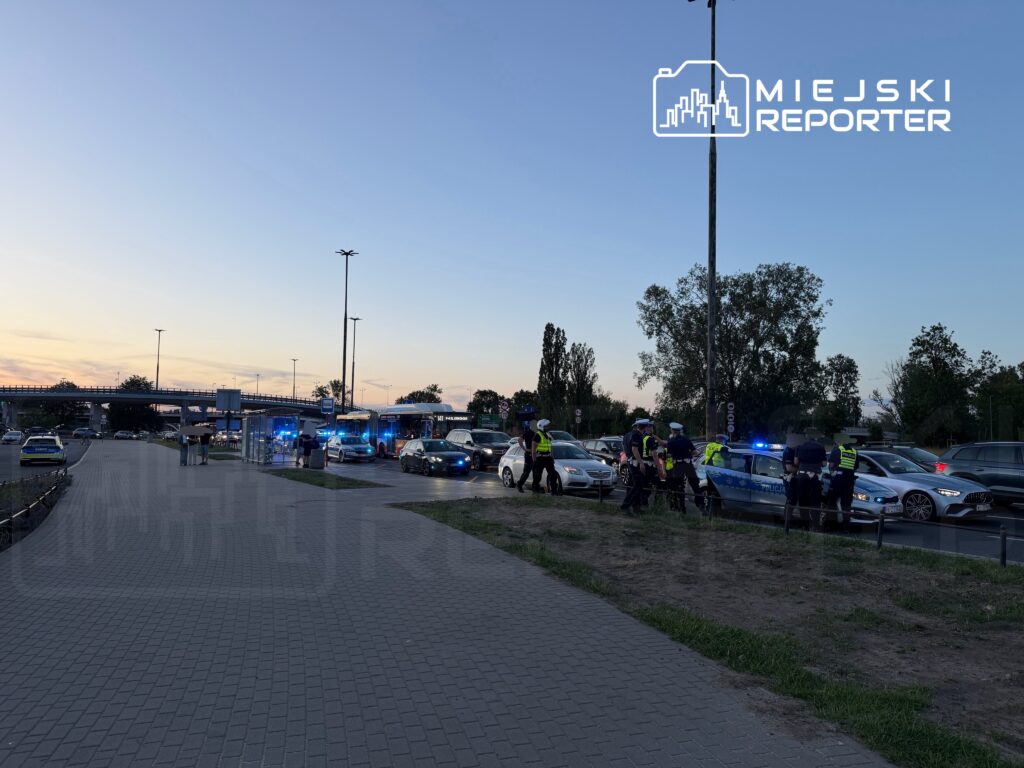 Grupa policjantów w mundurach prowadzi interwencję przy ruchliwej drodze, w tle widać zaparkowane samochody i radiowozy.