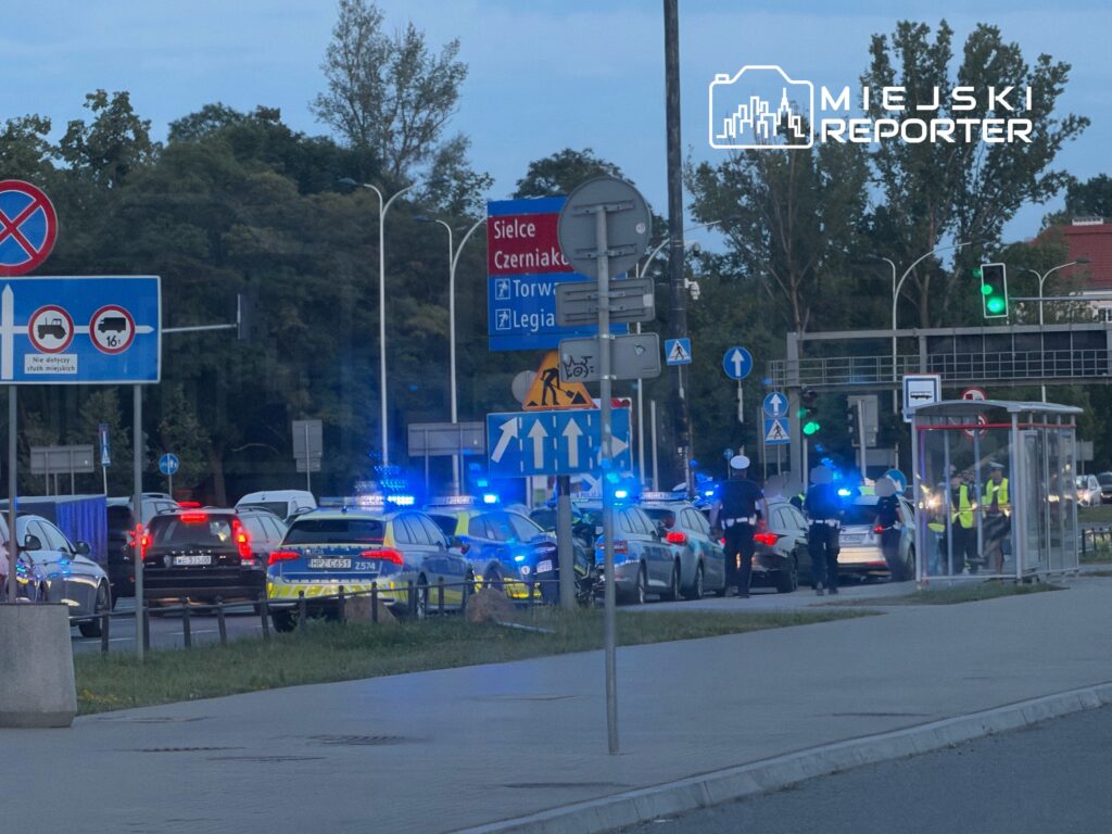 Ruch drogowy zatrzymany przez policję, w tle włączone sygnały świetlne radiowozów, znaki drogowe i przystanek autobusowy.