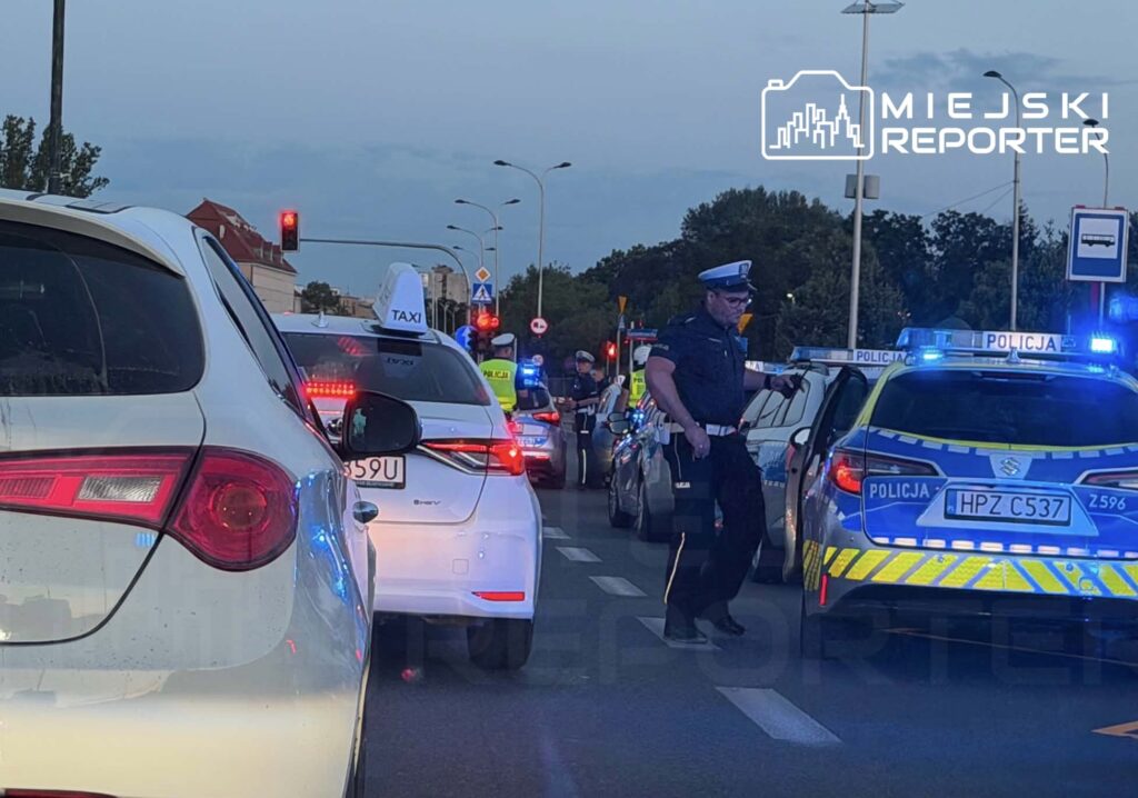 Policjant kontrolujący ruch drogowy na ulicy, obok zatrzymane samochody i radiowozy z włączonymi sygnałami świetlnymi.