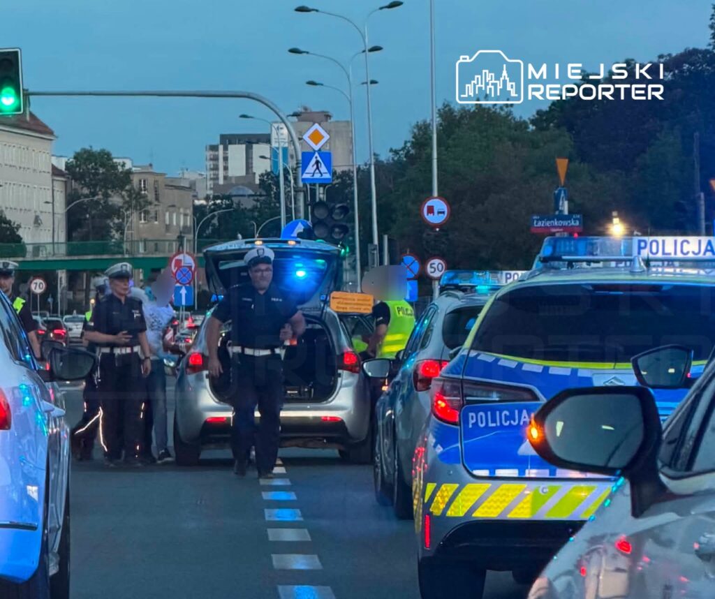 Grupa policjantów kontrolujących pojazdy na ruchliwej ulicy, w tle sygnalizacja świetlna i znaki drogowe.