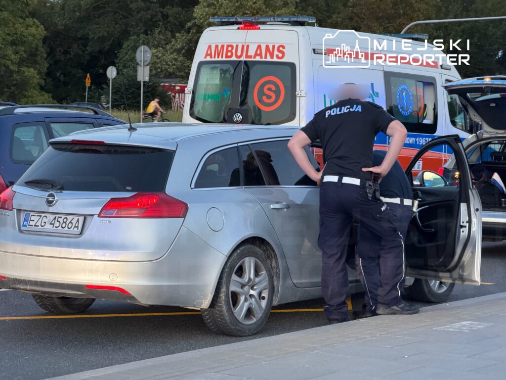 Policjanci sprawdzają wnętrze samochodu osobowego na drodze, obok zatrzymanego ambulansu.