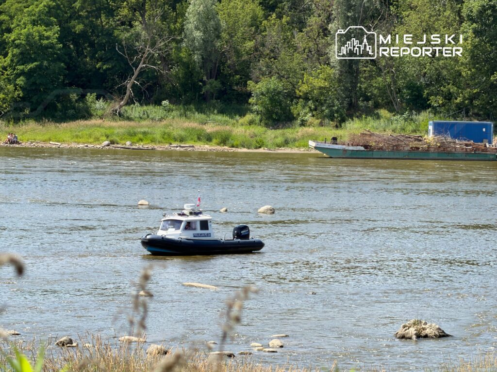 Mała łódź patrolowa przepływa wzdłuż rzeki, obok brzegu z zielonymi drzewami i grupą ludzi na plaży.
