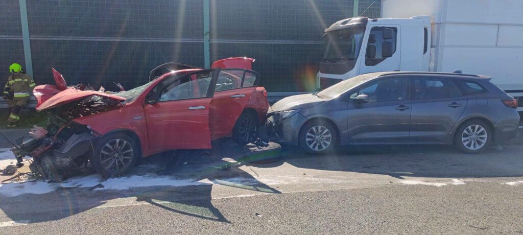 Czerwony samochód osobowy z uszkodzoną przednią częścią zderza się z szarym pojazdem na drodze, obok strażak w mundurze.