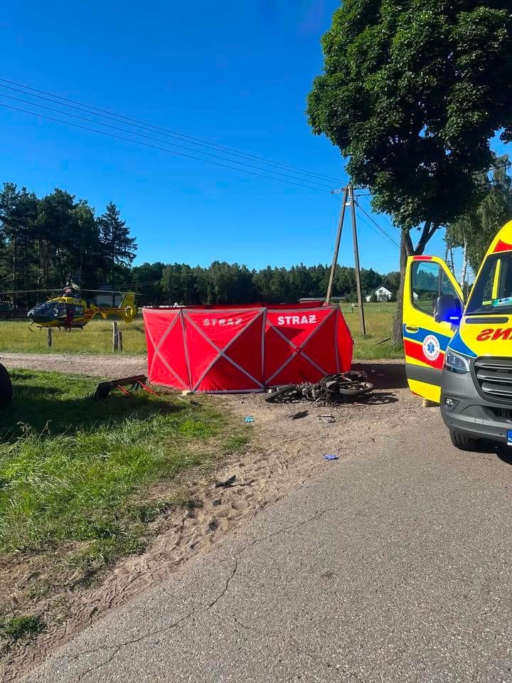 Na drodze stoi czerwony namiot ratunkowy z napisem "STRAŻ", obok zaparkowana karetka i śmigłowiec ratunkowy.