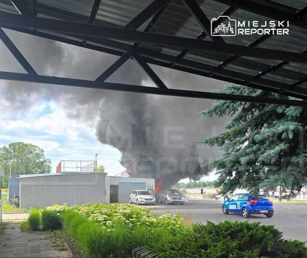Gęsty czarny dym unosi się nad zaparkowanymi samochodami obok budynku w strefie przemysłowej.