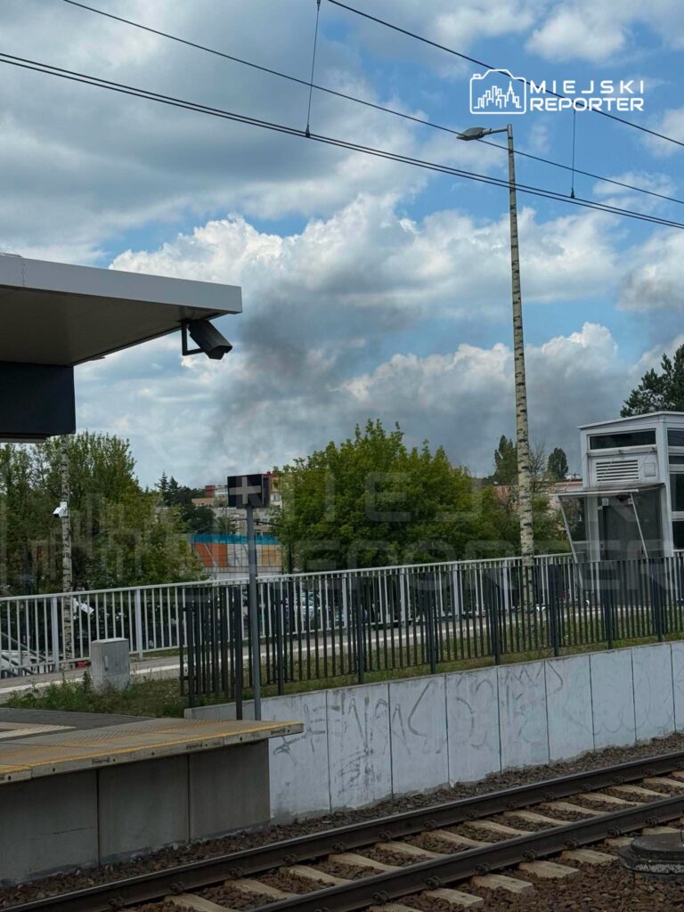 Ciemny dym unoszący się w tle nad stacją kolejową, w otoczeniu chmur i zieleni drzew.