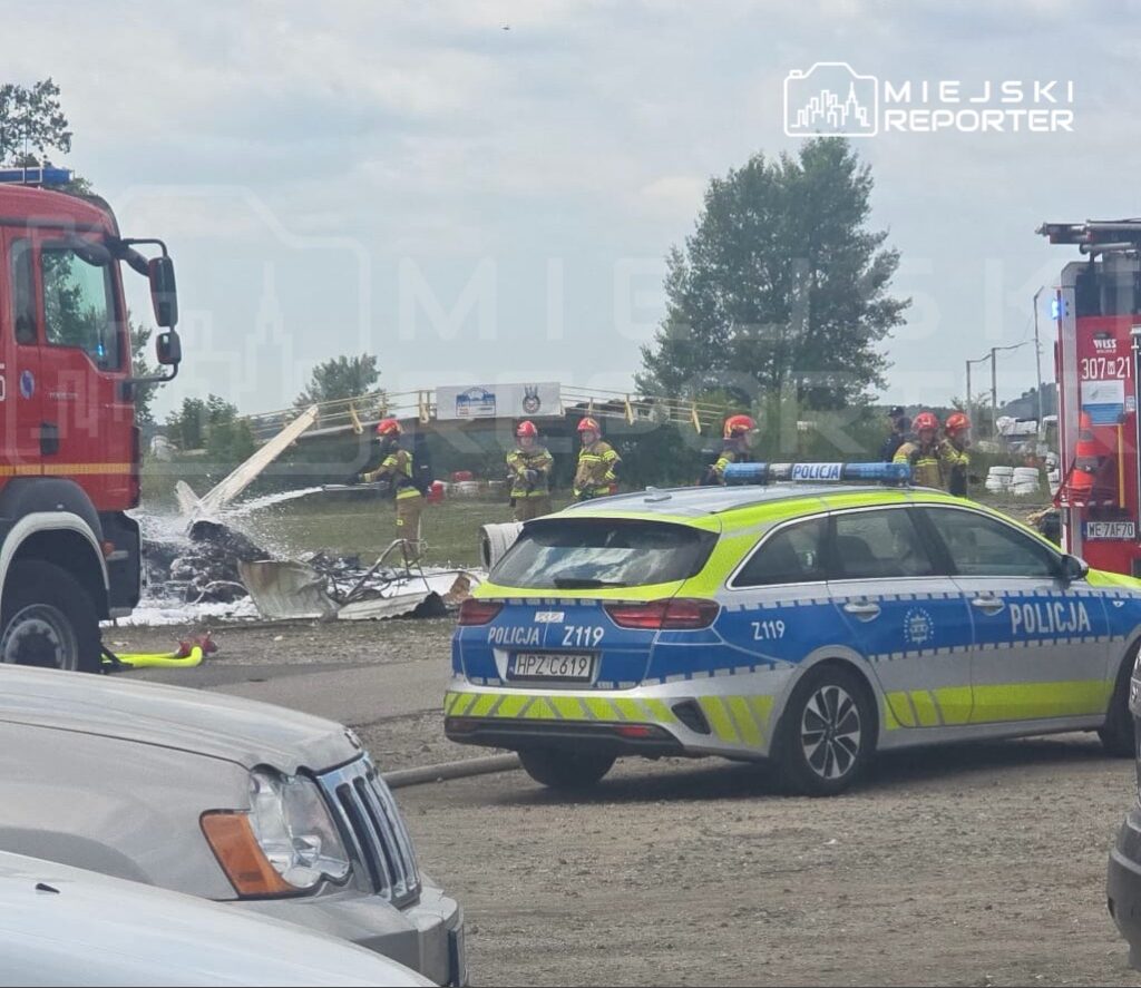 Strażacy w hełmach gaszą pożar wraku pojazdu na terenie akcji ratunkowej, obok stoi radiowóz policyjny.