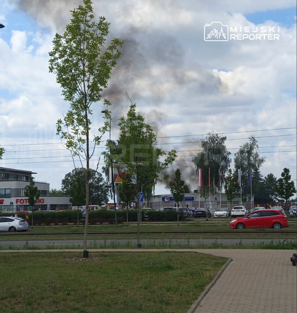 Czarny dym unoszący się z kierunku budynku w otoczeniu drzew i parkingu, w tle widoczne samochody.