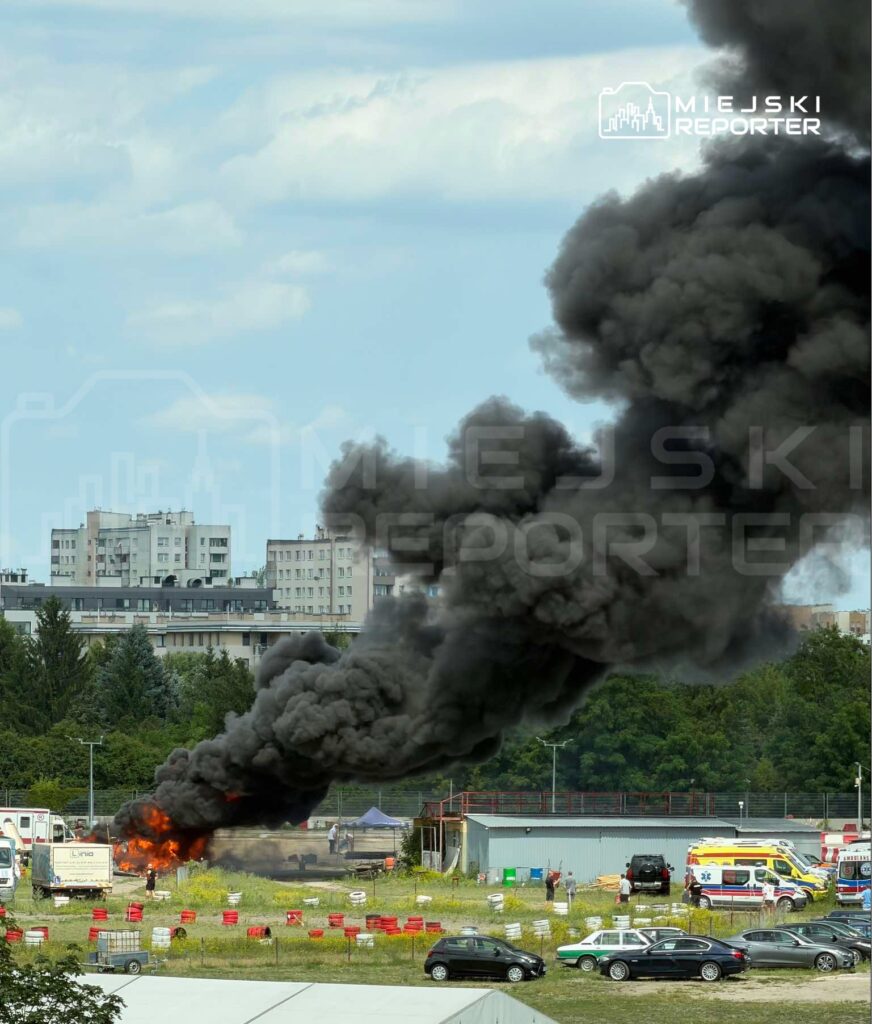 Czarny dym unosi się z płonącego pojazdu na terenie zielonym, w tle widać budynki mieszkalne oraz samochody.