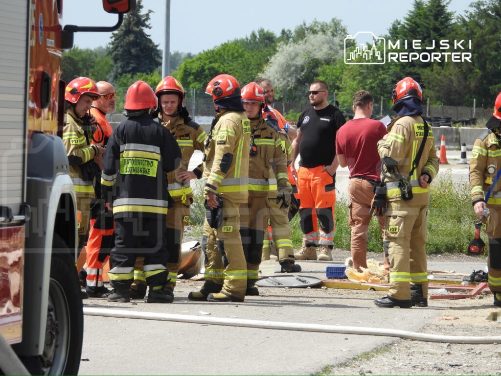 Grupa strażaków w hełmach omawia sytuację na miejscu zdarzenia, otoczona przez sprzęt ratunkowy i oznakowanie drogowe.