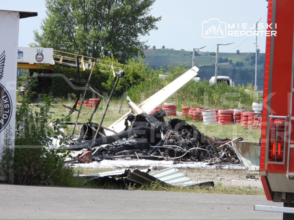 Wrak samolotu leżący na ziemi w zaroślach, otoczony przez szczątki i fragmenty konstrukcji, w tle widoczne bariery wyścigowe.