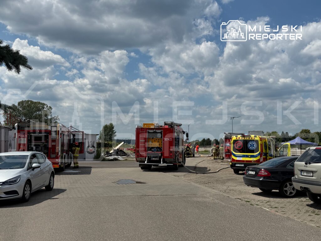 Wozy strażackie i ambulansy na terenie akcji ratunkowej, w tle zniszczony budynek pod chmurami.