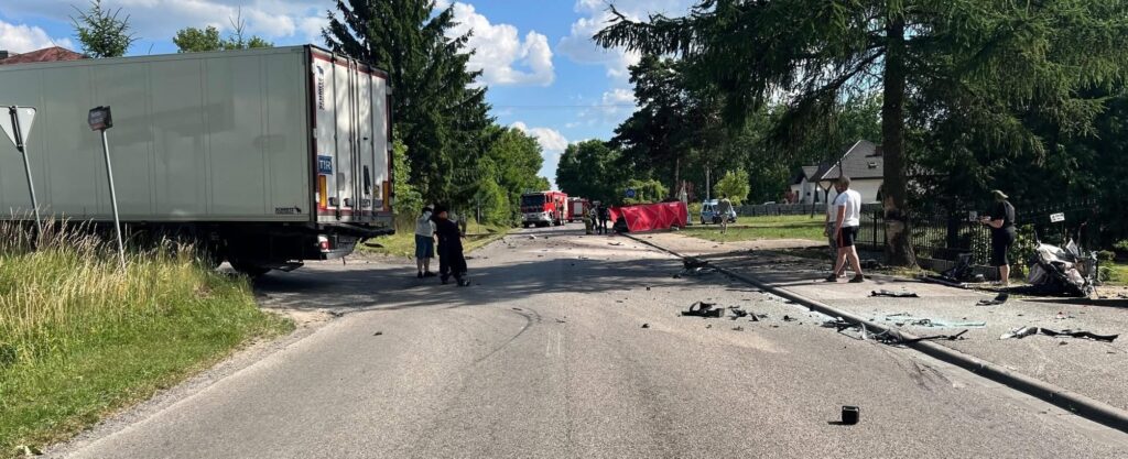 Na drodze leży rozbity samochód ciężarowy, a obok niego kilku ludzi oraz pojazdy służb ratunkowych.