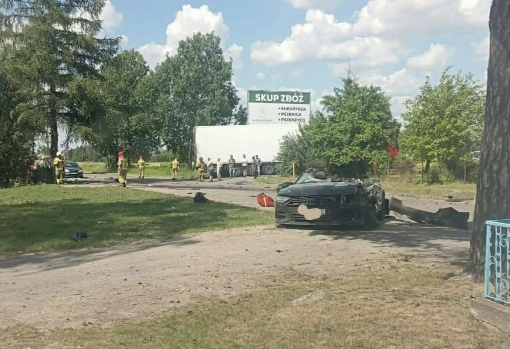 Na drodze leży przewrócone auto osobowe, a w tle grupa ratowników i pojazd ciężarowy przy sklepie rolnym.