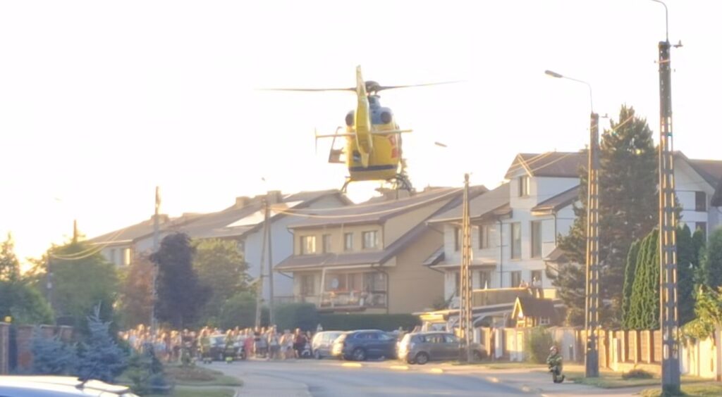 Helikopter ratunkowy unosi się nad osiedlem domów jednorodzinnych, a tłum ludzi obserwuje zdarzenie z ulicy.