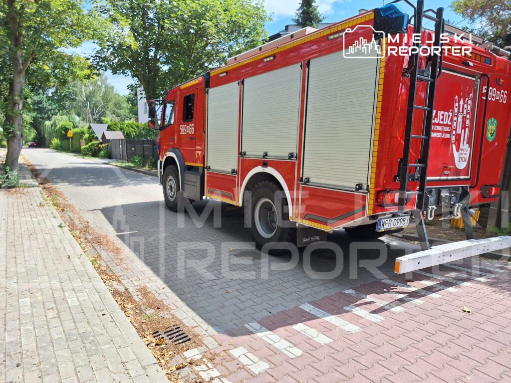 Czerwony wóz strażacki stoi na brukowanej drodze w otoczeniu drzew, obok widoczny znak drogowy.
