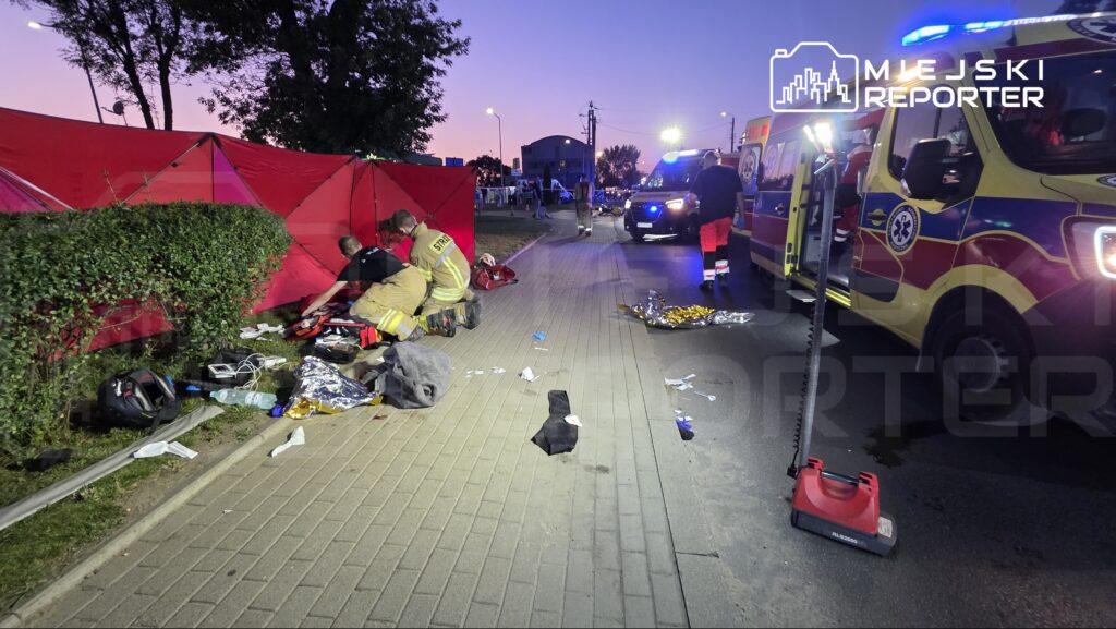 Strażacy udzielają pomocy poszkodowanym przy czerwonych namiotach na ulicy, obok ambulansów.
