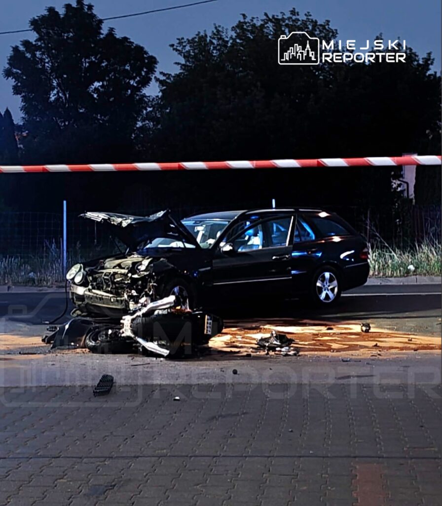 Uszkodzony samochód osobowy z otwartą maską leży na drodze, obok widać fragmenty wraku motocykla.