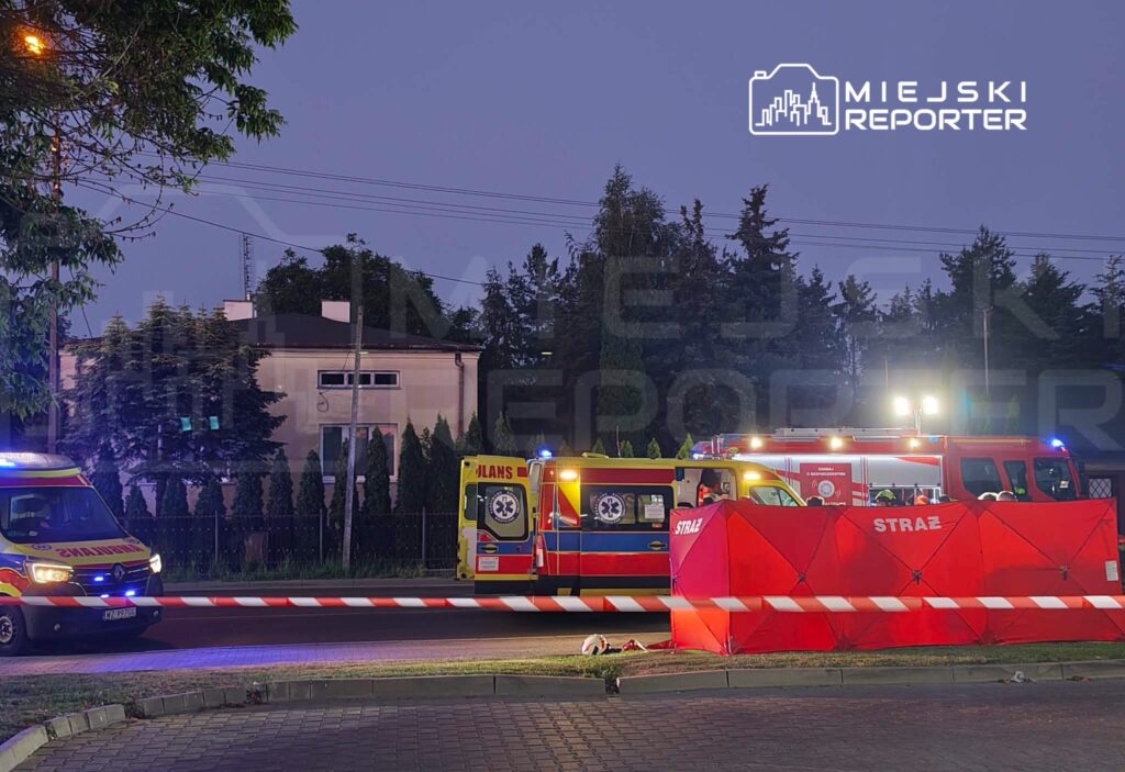 Karetka pogotowia i wóz strażacki na miejscu zdarzenia, otoczone czerwonymi barierkami w wieczornym świetle.