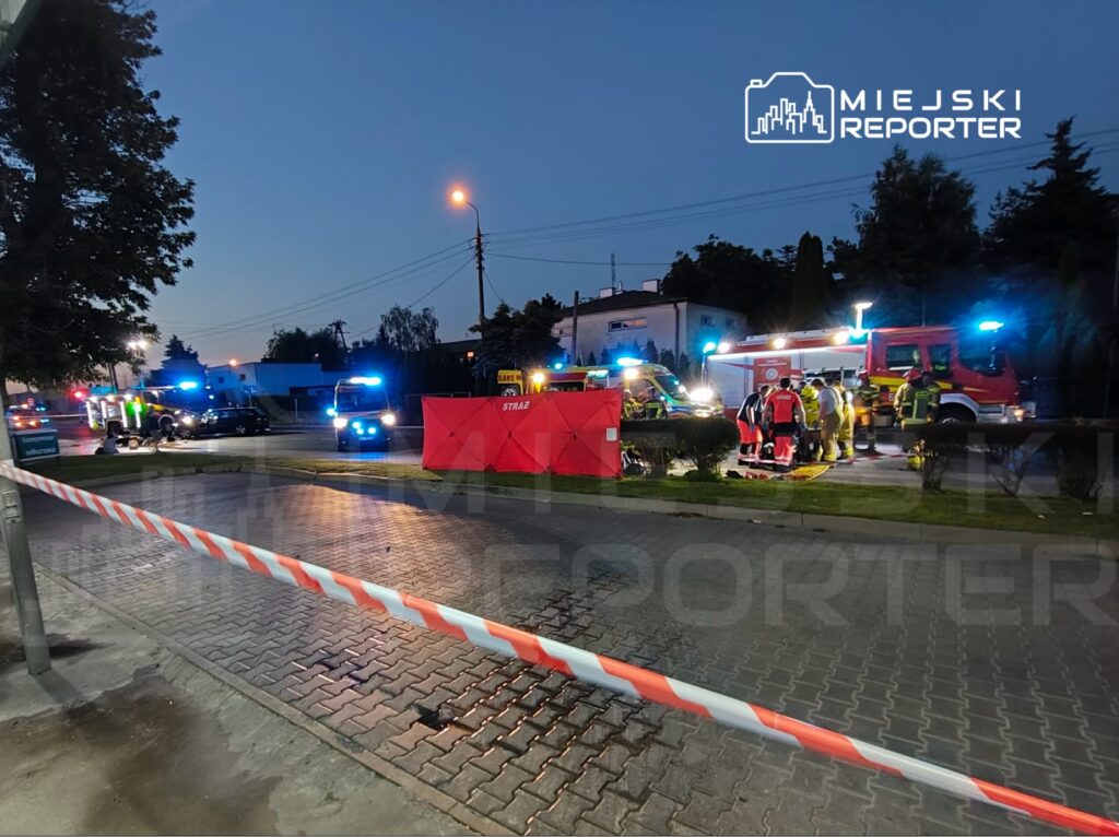 Grupa ratowników medycznych i strażaków pracuje na miejscu zdarzenia w otoczeniu pojazdów ratunkowych oraz policyjnych taśm.