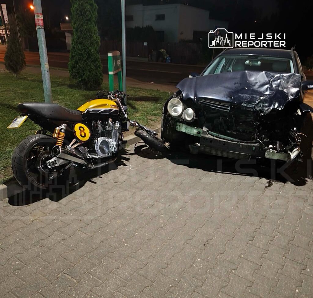 Motocykl z żółtymi detalami stoi obok uszkodzonego samochodu osobowego na oświetlonym parkingu.