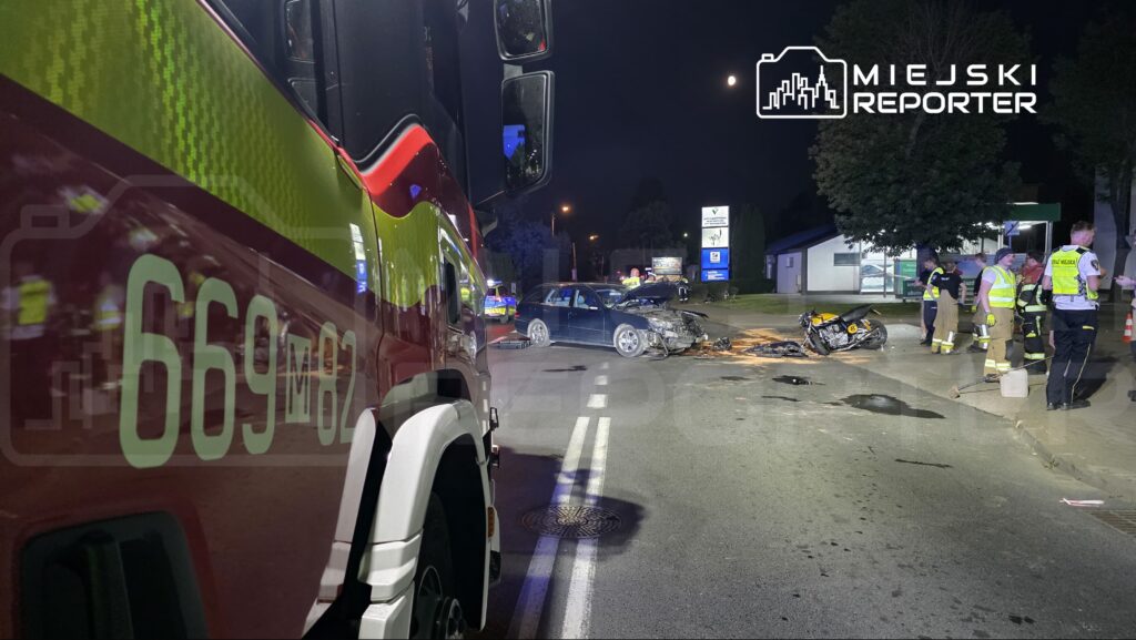 W nocy na drodze doszło do wypadku; uszkodzony samochód osobowy i motocykl, obok pracujący ratownicy medyczni.