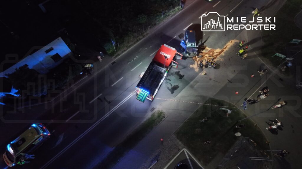 W nocy widać wypadek drogowy, w którym uczestniczy czerwona ciężarówka i niebieski pojazd osobowy, na jezdni leżą resztki ładunku. Obok pracują służby ratunkowe, a w tle zebrani są ciekawscy.