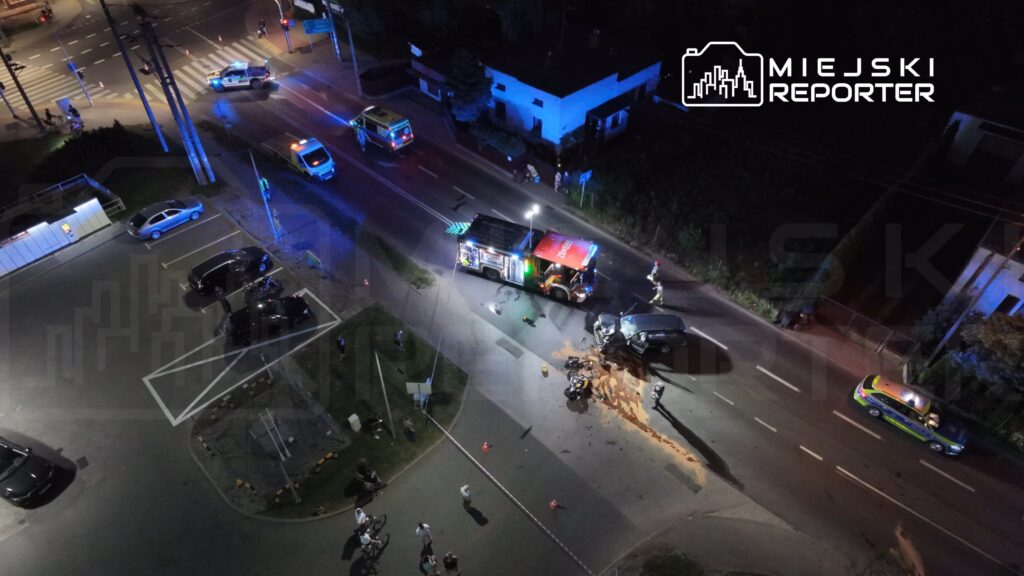Na drodze leży uszkodzony samochód, a obok niego znajdują się strażacy i policjanci. W tle widać radiowozy i karetki.