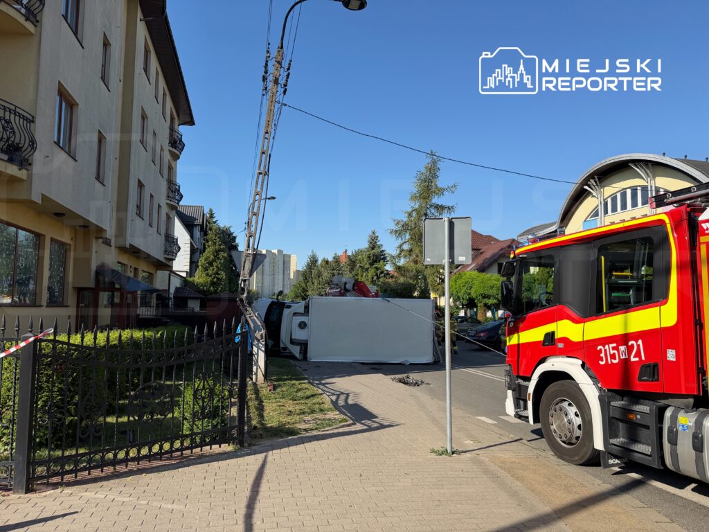 Straż pożarna przy przewróconym pojeździe na ulicy, obok budynków mieszkalnych i drzew.