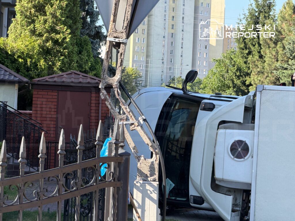 Pojazd dostawczy leży na boku obok ogrodzenia, w tle widać zielone drzewa i budynki mieszkalne.