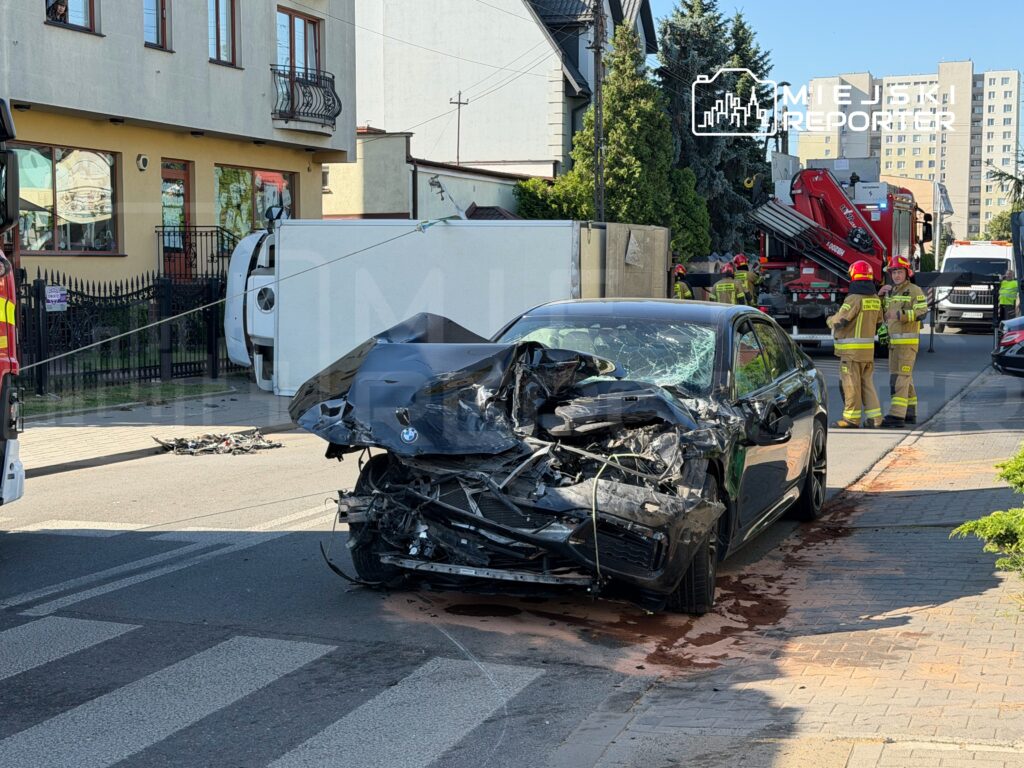 Zniszczony czarny samochód osobowy stoi na drodze, obok przewrócony biały pojazd, a w tle pracują strażacy.