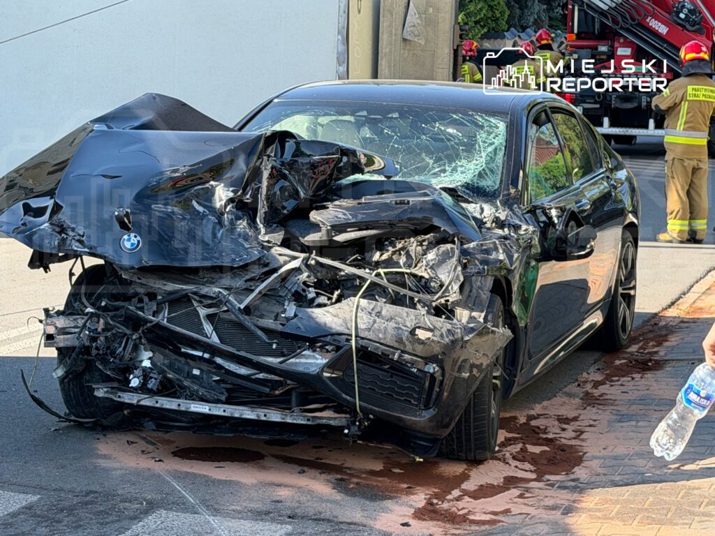 Zniszczony samochód osobowy z uszkodzoną karoserią stoi na ulicy, obok pracują strażacy.