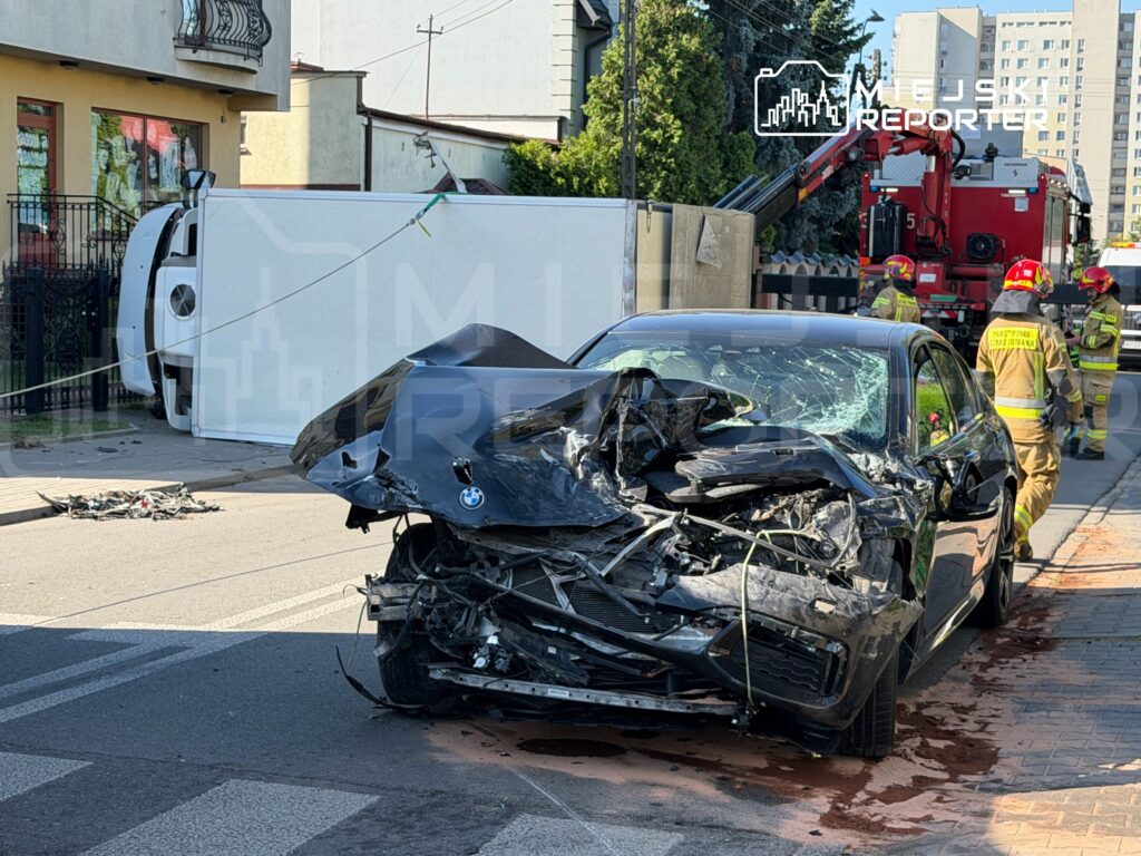 Zniszczony samochód osobowy leży na jezdni obok przewróconego furgonu, a strażacy pracują nad usunięciem wraku.