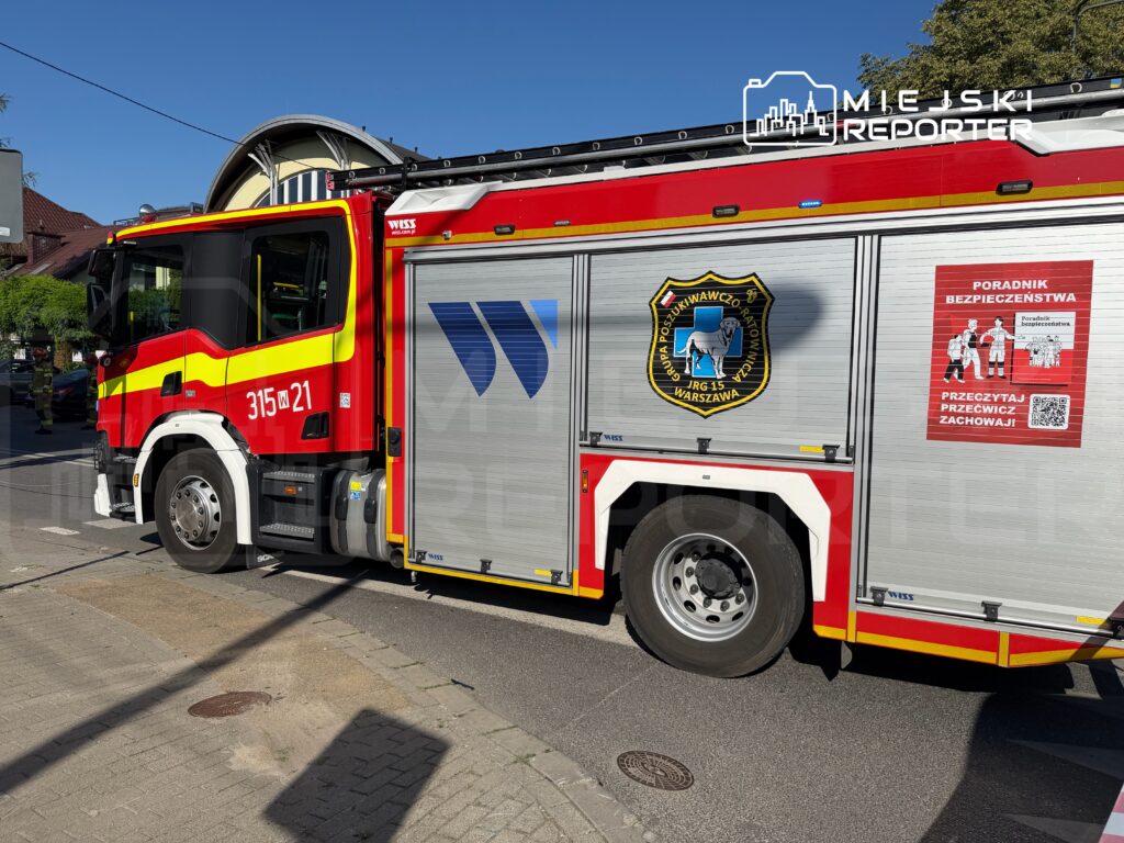 Czerwony wóz strażacki z numerem 315 w Warszawie stoi na ulicy obok budynku, a na drzwiach widnieje herb jednostki.