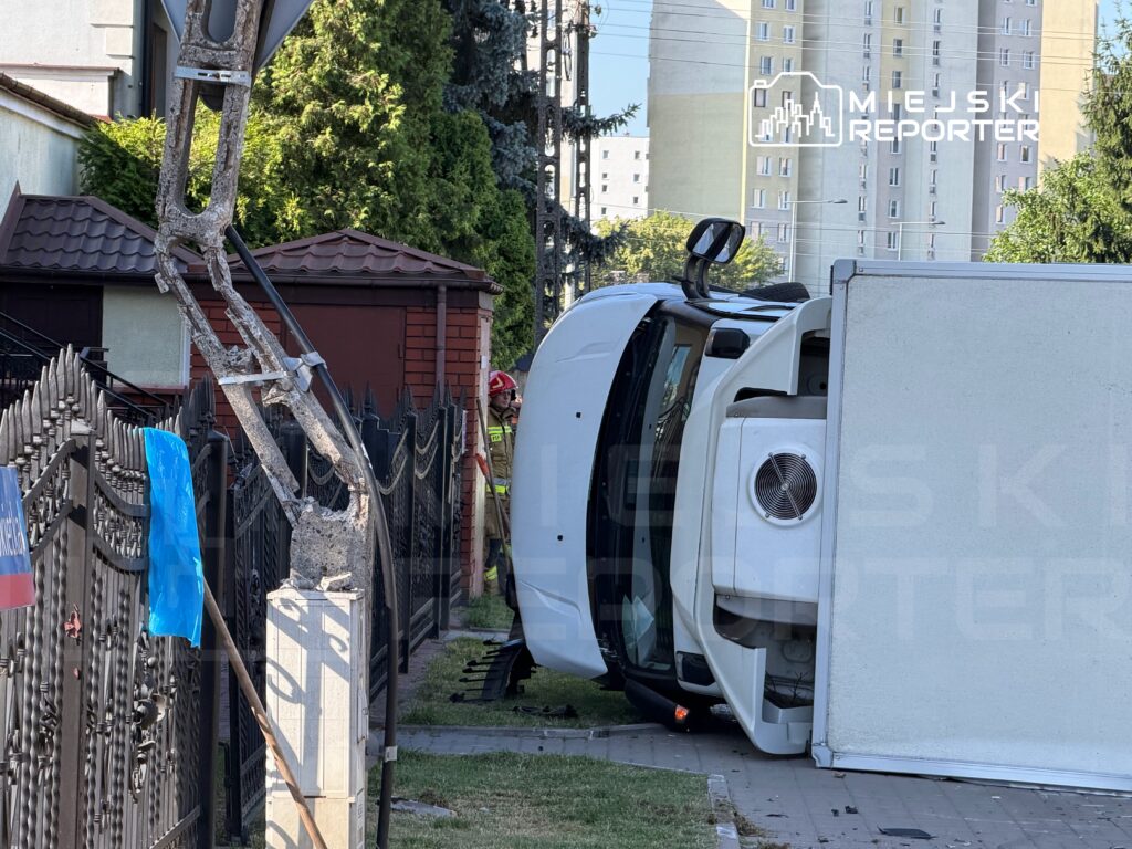 Samochód dostawczy leży na boku na trawniku obok ogrodzenia, a strażak stoi w pobliżu, obserwując sytuację.