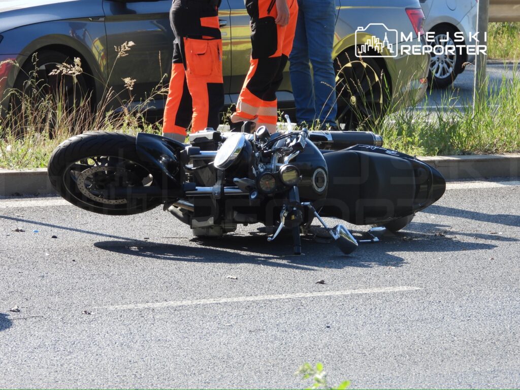 Motocykl leży na boku na jezdni, a obok niego stoją osoby w pomarańczowych kamizelkach odblaskowych.