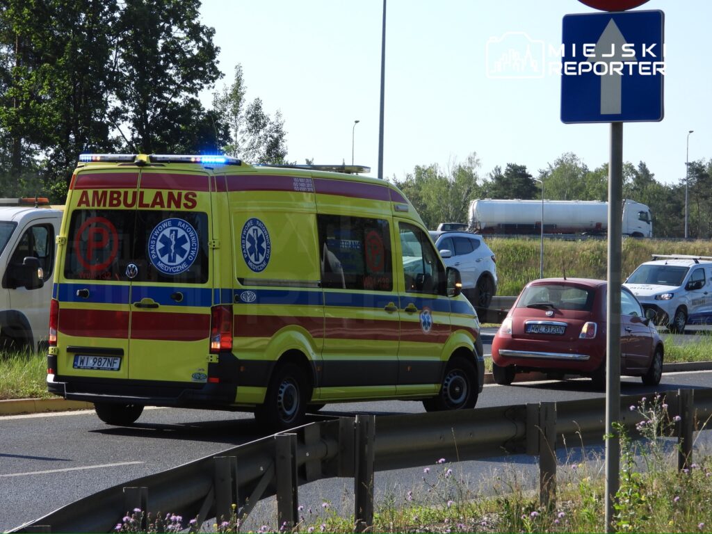 Karetka pogotowia z włączonymi sygnałami świetlnymi zatrzymana na drodze, obok innych pojazdów w tle.