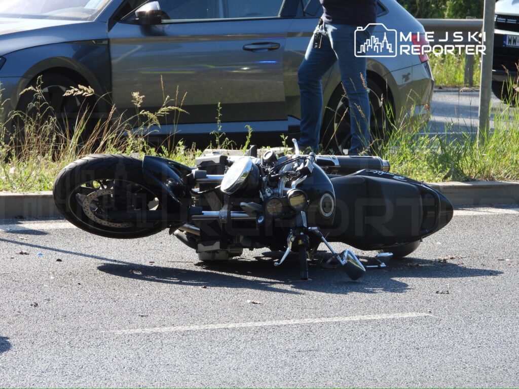 Motocykl leżący na boku na jezdni, obok stoi mężczyzna z kluczami w ręku, w tle widać zaparkowane samochody.