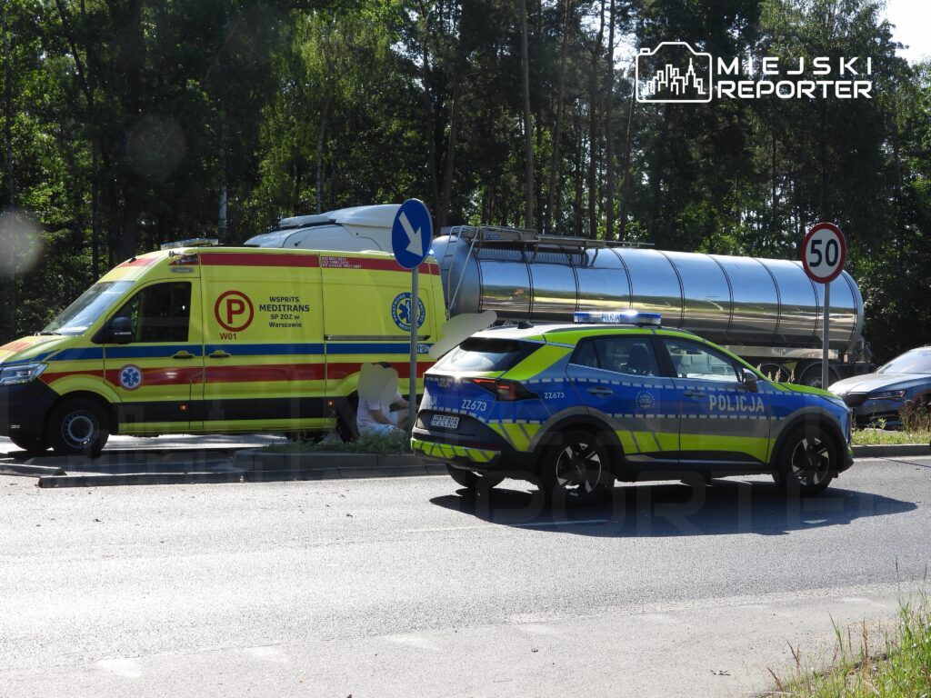 Karetka pogotowia i radiowóz policyjny zatrzymane przy drodze, obok dużego pojazdu transportowego, w otoczeniu zieleni.