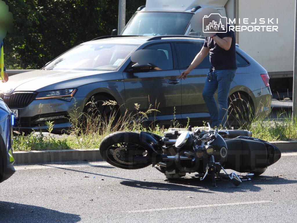 Motocykl leży na boku na jezdni, a mężczyzna w niebieskiej koszulce rozmawia przez telefon obok.