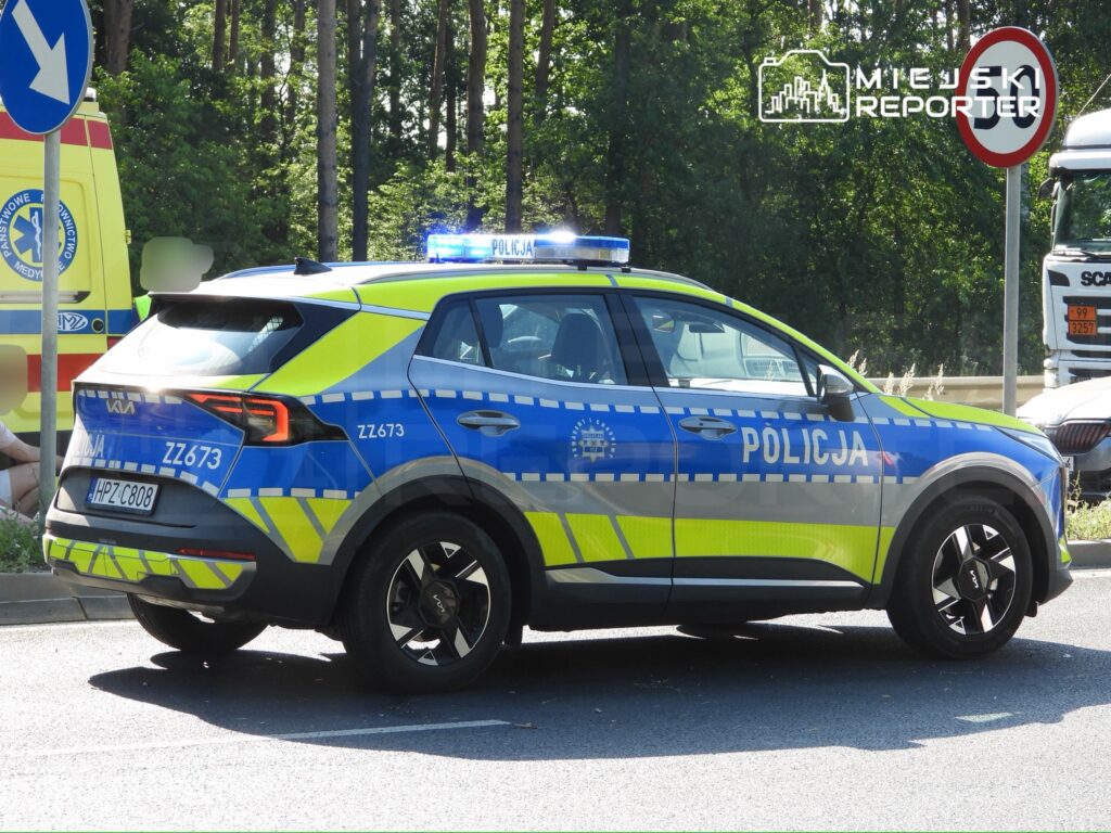 Policyjny radiowóz z niebieskimi światłami zatrzymany przy drodze, obok karetki pogotowia.
