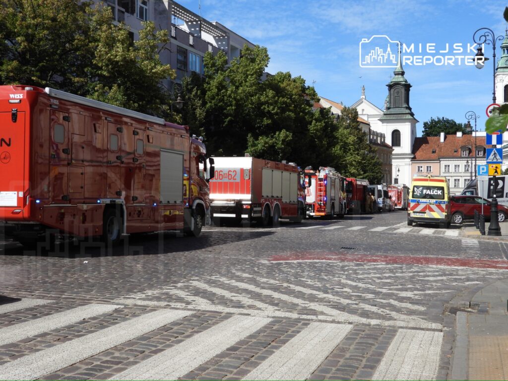Rząd pojazdów strażackich zaparkowanych wzdłuż brukowanej ulicy, w tle widoczny kościół i zielone drzewa.