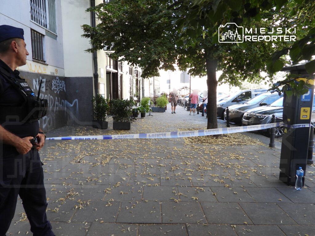 Policjant w mundurze stoi przy taśmie zabezpieczającej na ulicy, obok parkujących samochodów i opadających liści.
