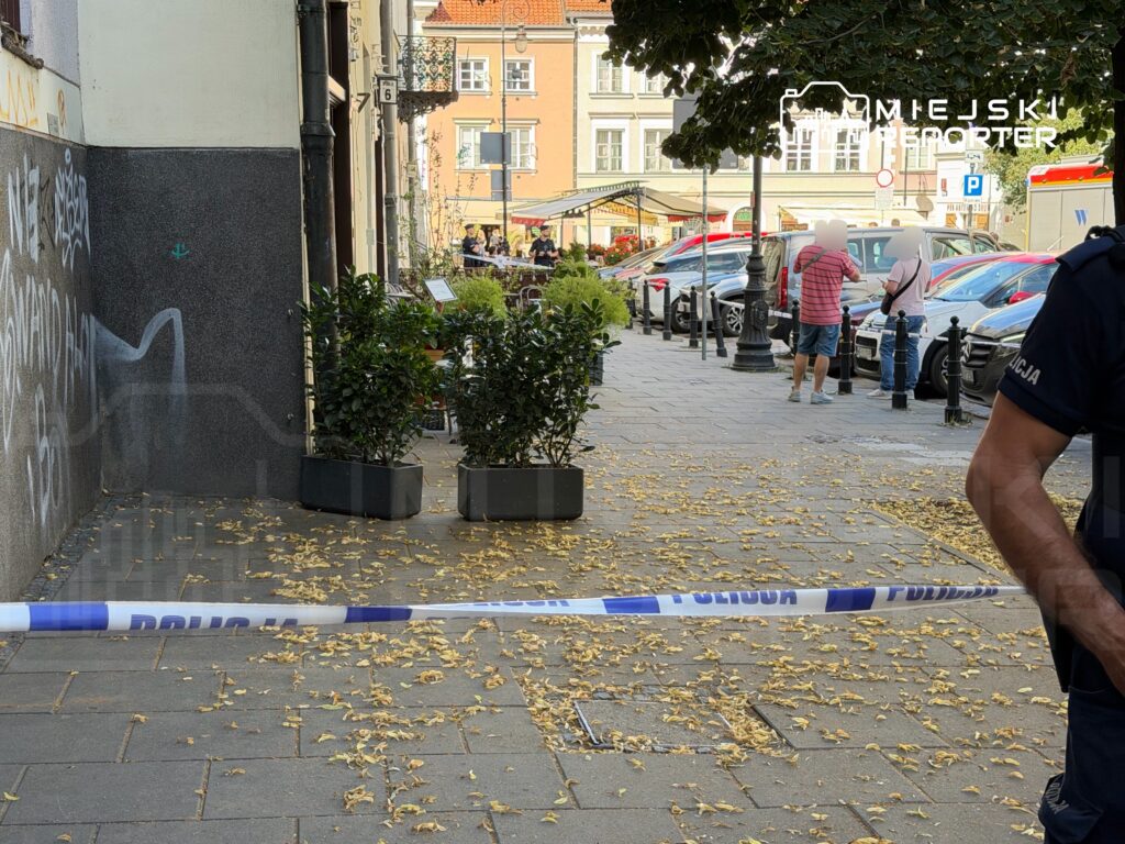 Policjant stojący przy taśmie zabezpieczającej, a w tle przechodnie rozmawiający na ulicy pokrytej opadłymi liśćmi.