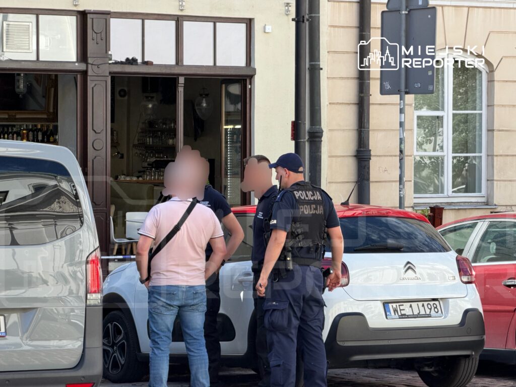 Mężczyzna w białej koszulce rozmawia z policjantami na ulicy przed lokalem gastronomicznym.