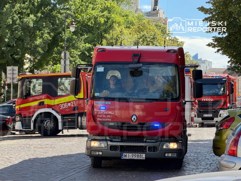 Czerwony wóz strażacki z włączonymi sygnałami świetlnymi przemieszcza się przez ulicę, obok innego wozu strażackiego.