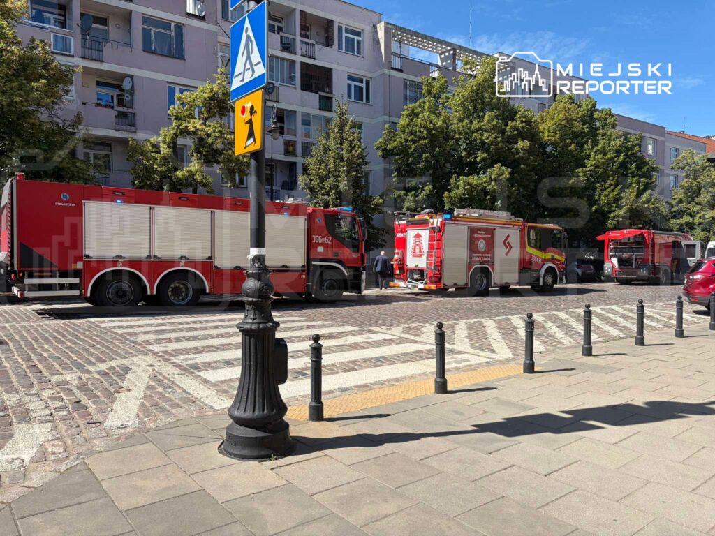 Czerwone samochody strażackie zaparkowane na ulicy w pobliżu budynków mieszkalnych, otoczone drzewami.